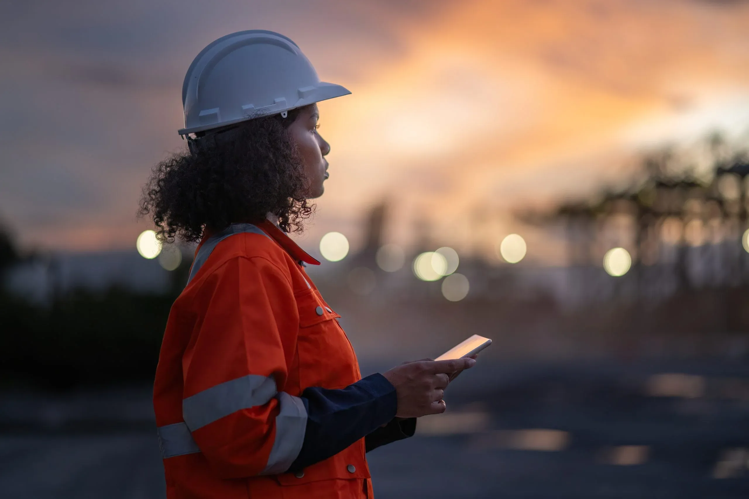 Worker in a white hard hat and orange high-visibility jacket holding a tablet at dusk